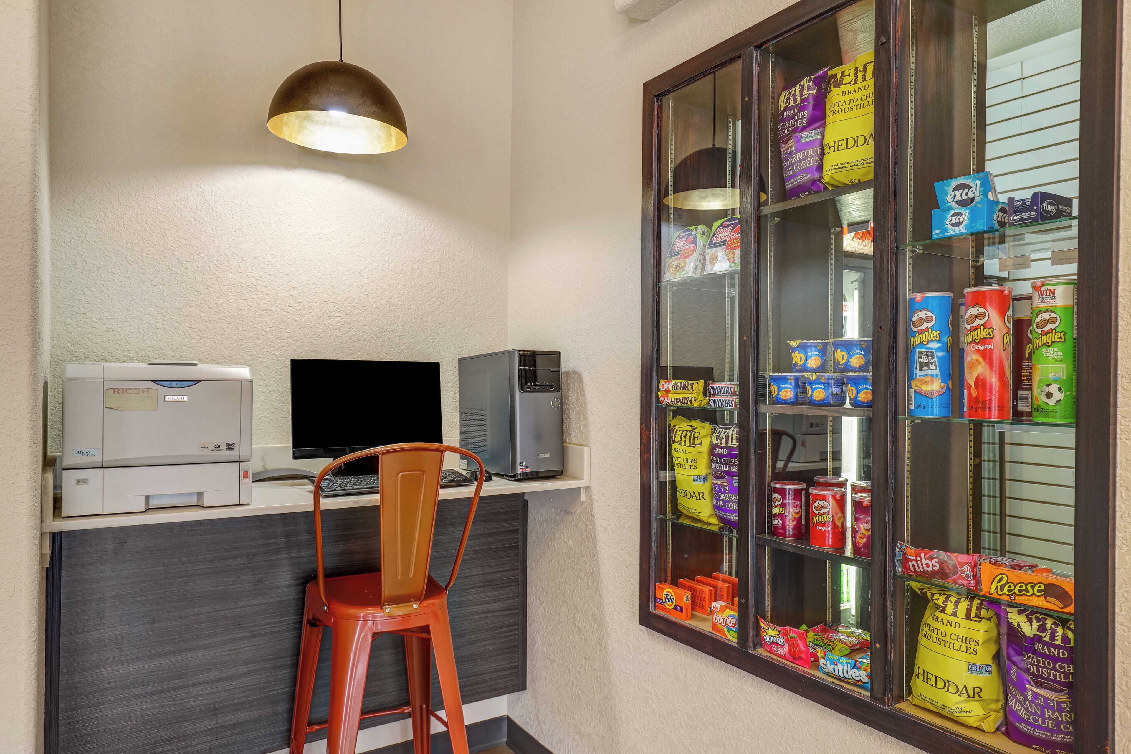 A small office with a desk, chair, printer, computer, and a snack vending machine.