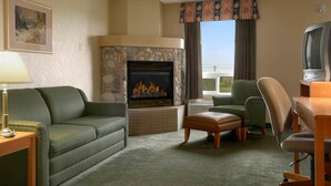 A hotel room with a stone fireplace, green sofa, armchair, ottoman, and a television on a wooden table.
