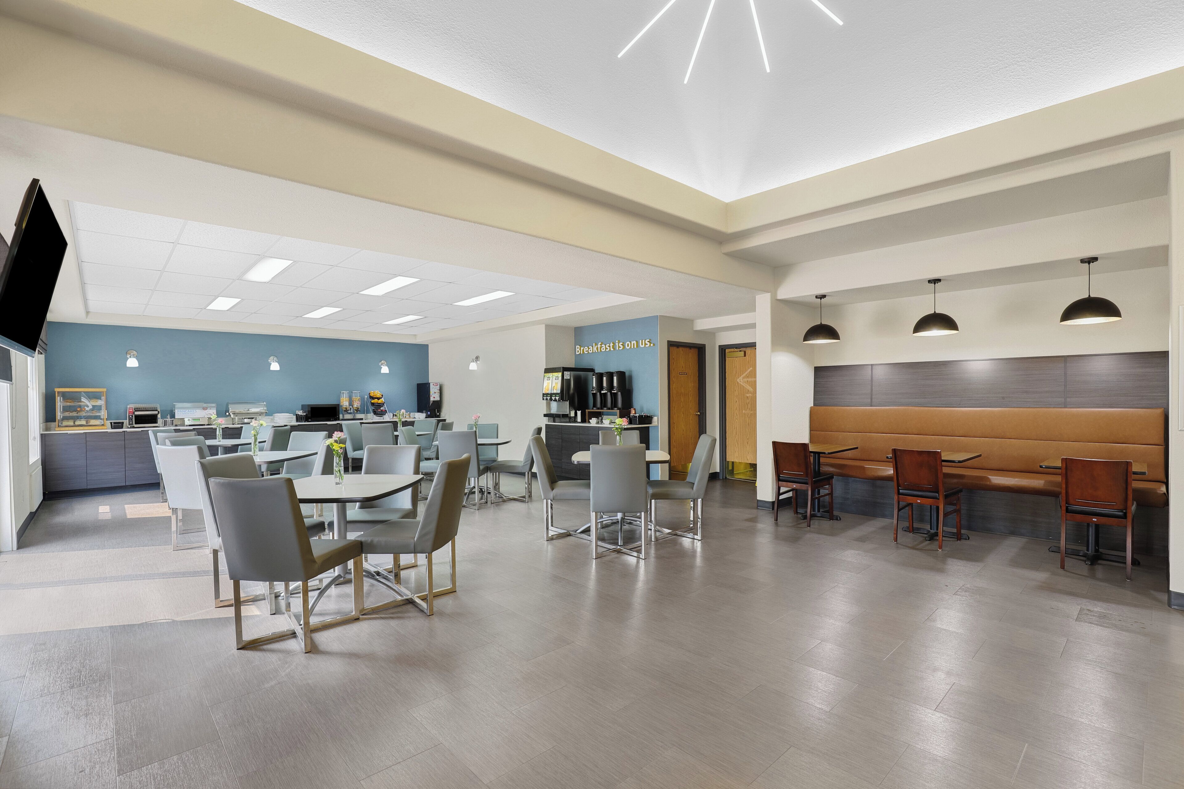 A modern dining area with tables and chairs, a counter with a coffee machine, and a sign that reads 'breakfast is on us.'