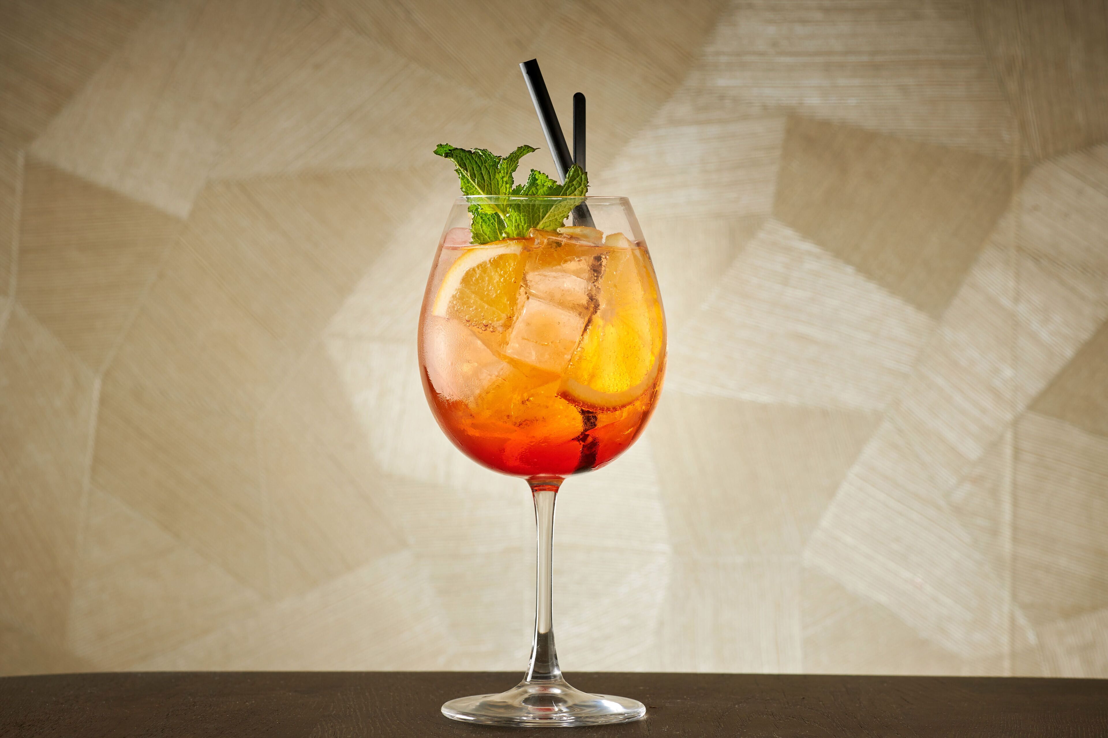 A cocktail glass with ice cubes, a slice of orange, and a mint garnish on a wooden table.