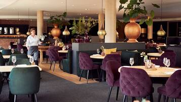 Un restaurant avec des chaises recouvertes d’un tissu violet, un coin bar et de grandes plantes en pot.
