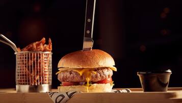 Ein Burger mit Messer darauf, eine Pommes-Korb und eine Tasse Kaffee auf einem Holztisch.
