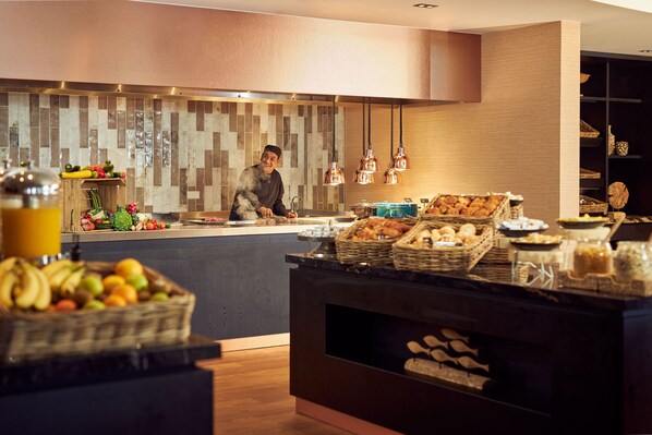 A modern kitchen with a chef preparing food, a fruit display, and a buffet area with various dishes. - Van der Valk Hotel Leusden - Amersfoort (Leusden-Centrum)
