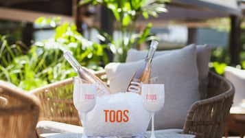 A wicker chair with a white cushion, a small round table, and a bucket filled with ice and two champagne flutes labeled "TOOS".