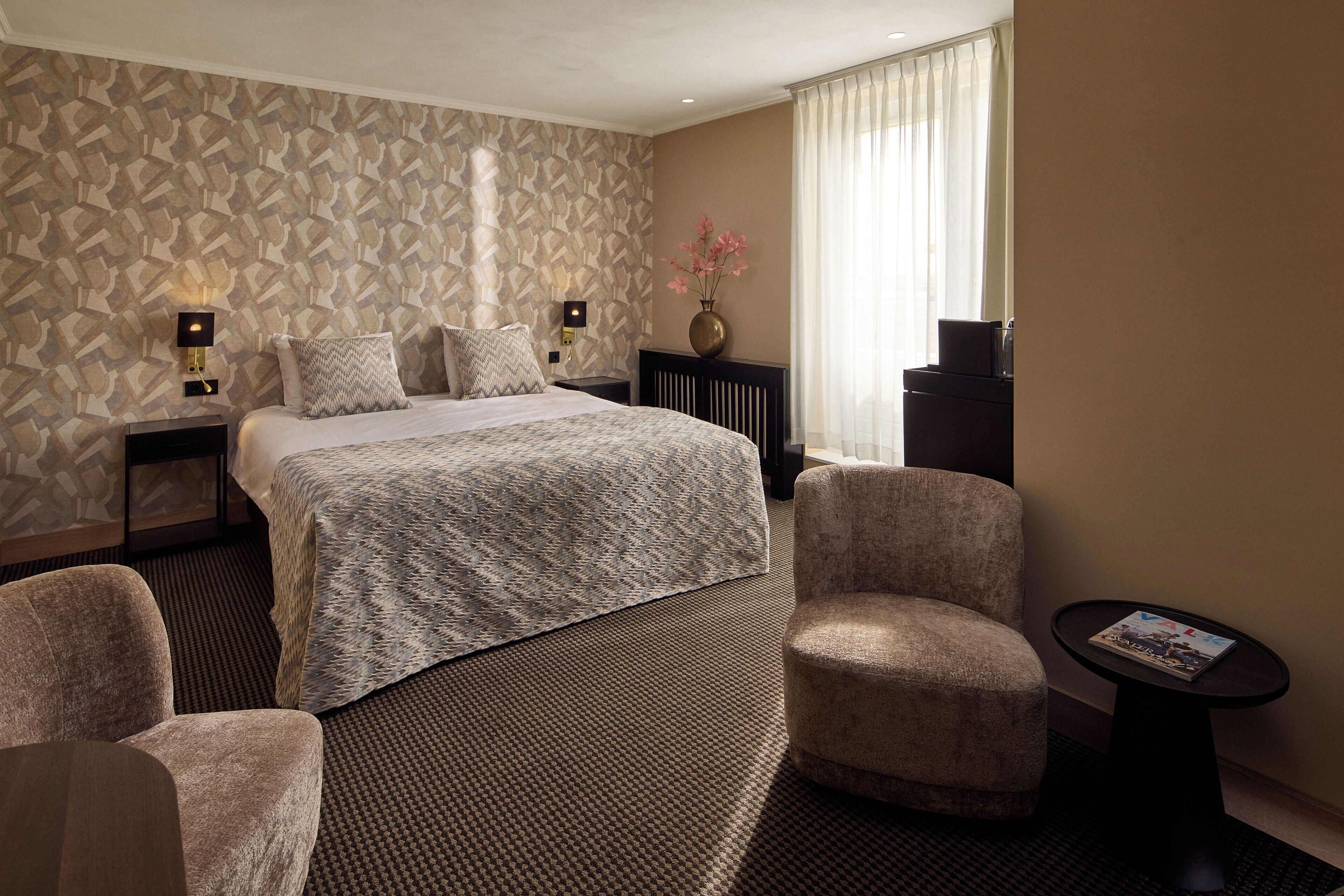 A hotel room with a large bed, two bedside tables, a chair, a small table, and a vase with flowers.