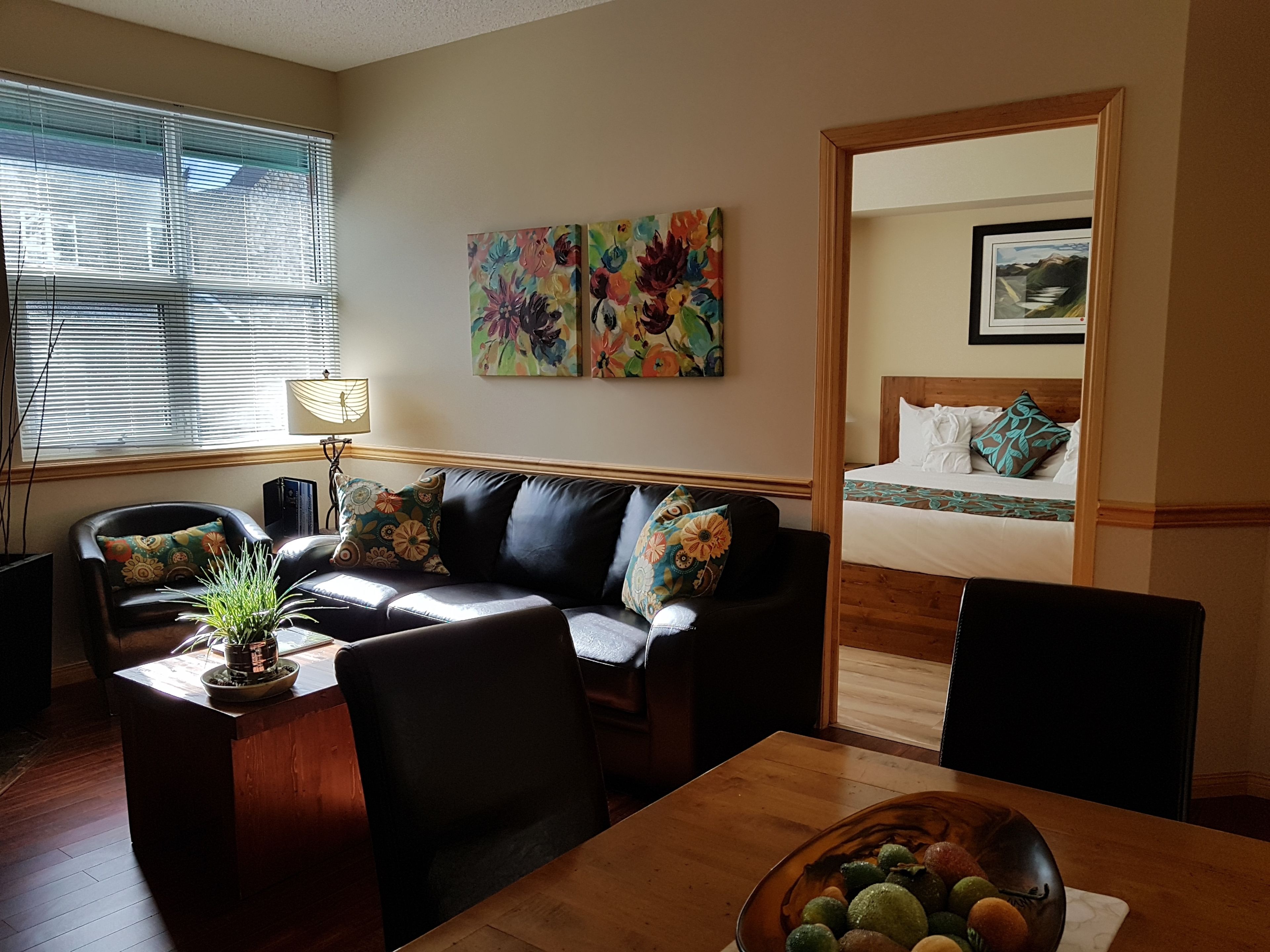 A living room with a sofa, a dining table, and a small kitchen area. There is a bed in the adjacent room.