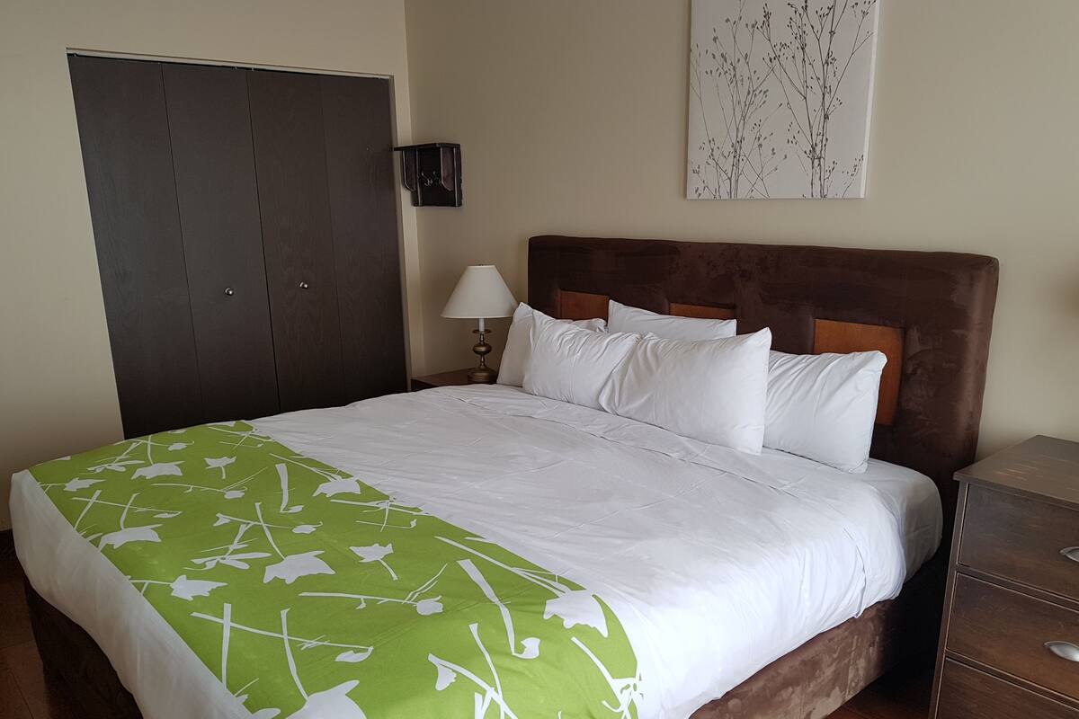 A neatly made bed with white bedding and a green floral bedspread, a wooden headboard, a bedside lamp, a wooden nightstand, and a wardrobe.