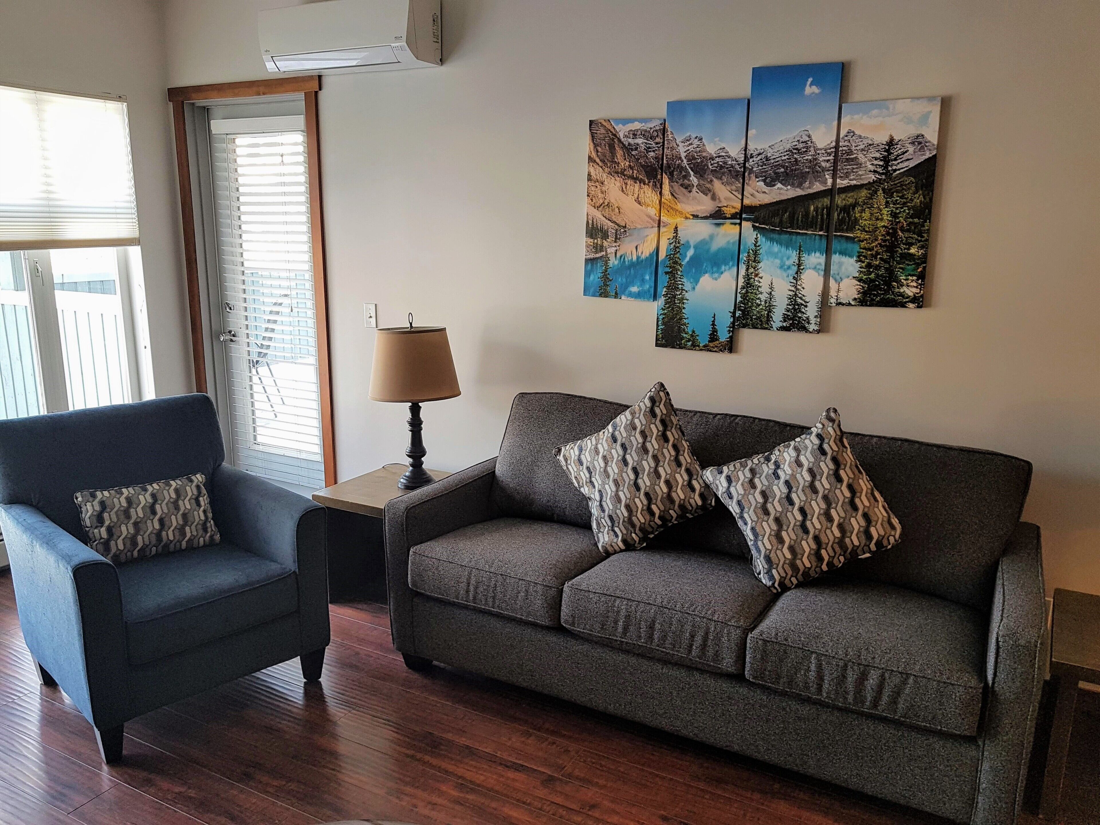 Ein Wohnzimmer mit grauem Sofa, blauem Sessel und gemusterten Kissen. Auf einem Holztisch steht eine Lampe, und ein Fenster mit Jalousien ist zu sehen.