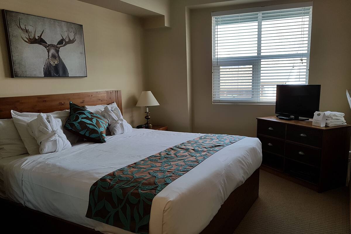 A neatly made bed with a patterned bedspread, a wooden headboard, and a painting of a moose on the wall.