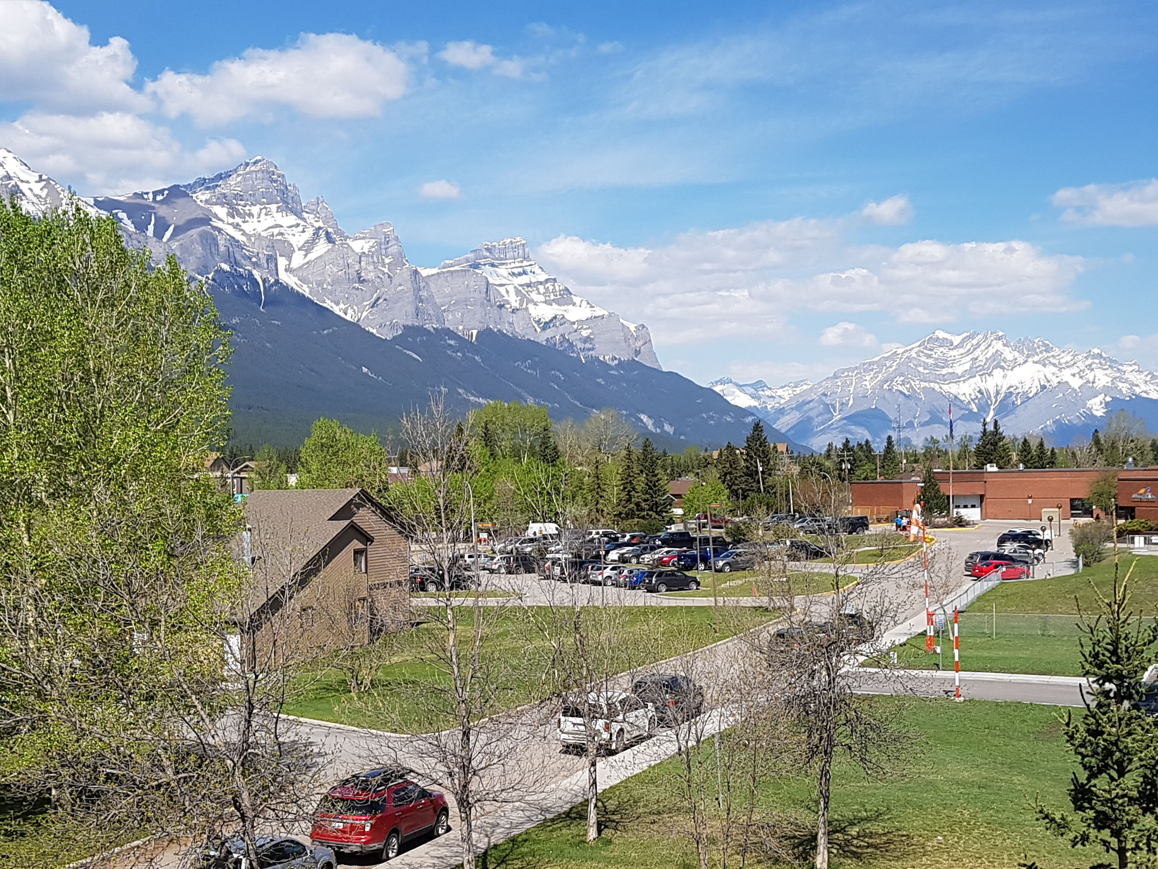 Un parking avec des voitures et un bâtiment au premier plan, et des montagnes enneigées en arrière-plan.