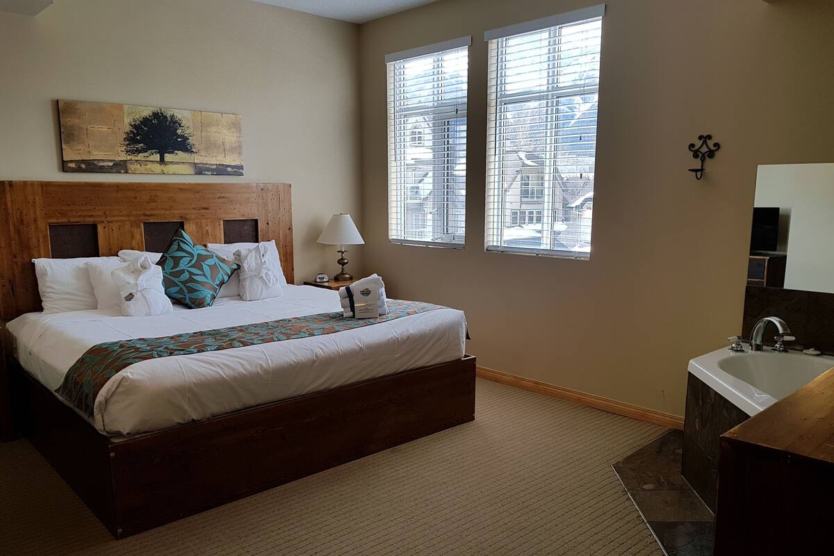 A hotel room with a large bed, a wooden headboard, a painting of a tree, two windows with blinds, a bathroom with a sink and mirror, and a carpeted floor.