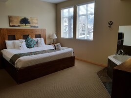 Quarto de hotel com uma cama grande, cabeceira de madeira, pintura de uma árvore, duas janelas com persianas, banheiro com pia e espelho, e piso coberto por carpete.