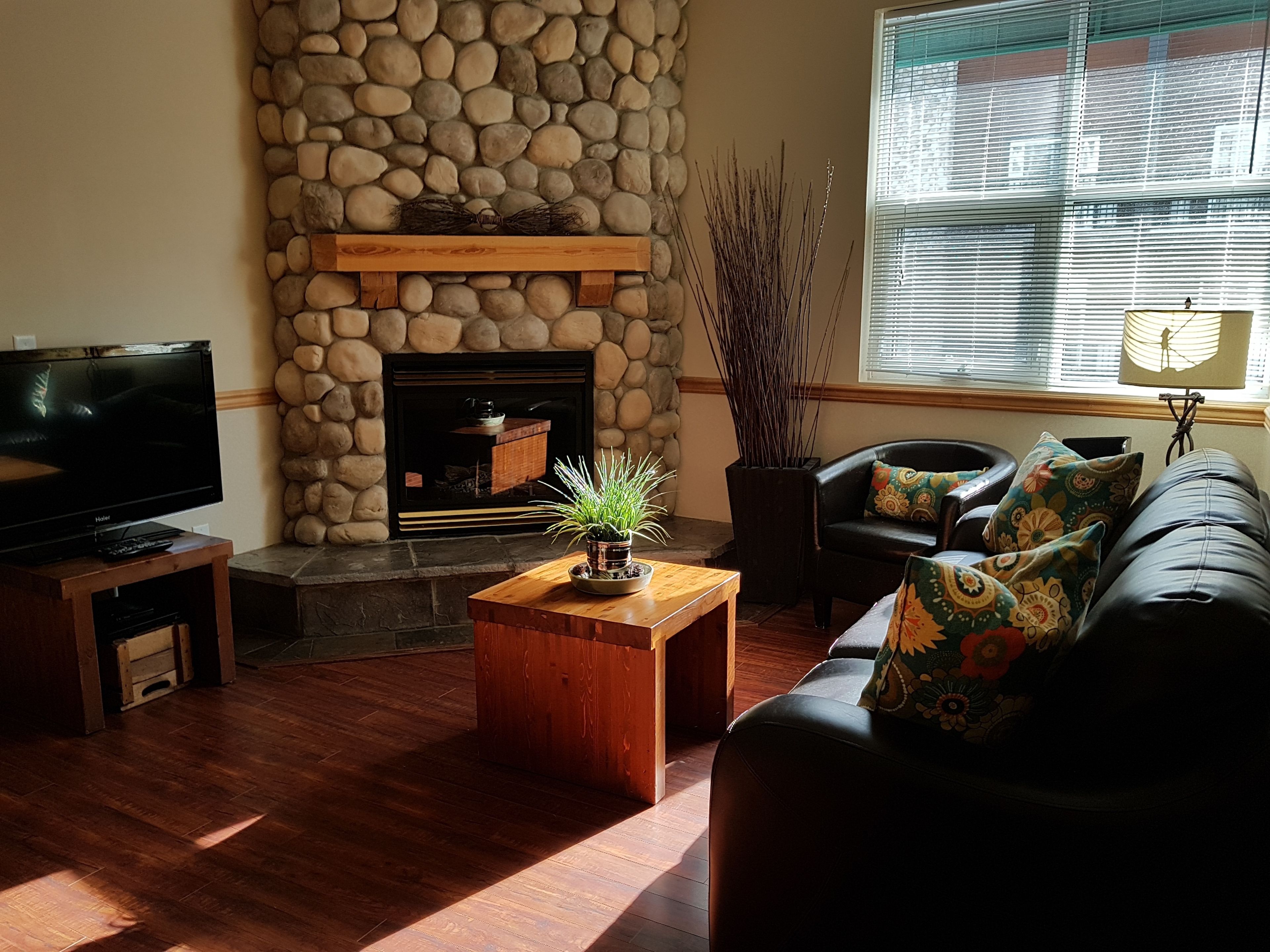 A living room with a stone fireplace, a television, a sofa with patterned cushions, and a window with blinds.