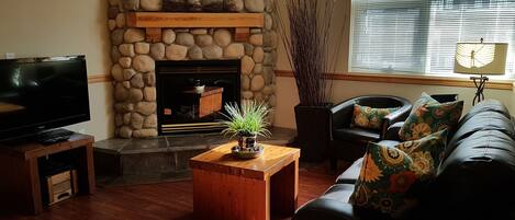 A living room with a stone fireplace, a television, a sofa with patterned cushions, and a window with blinds.