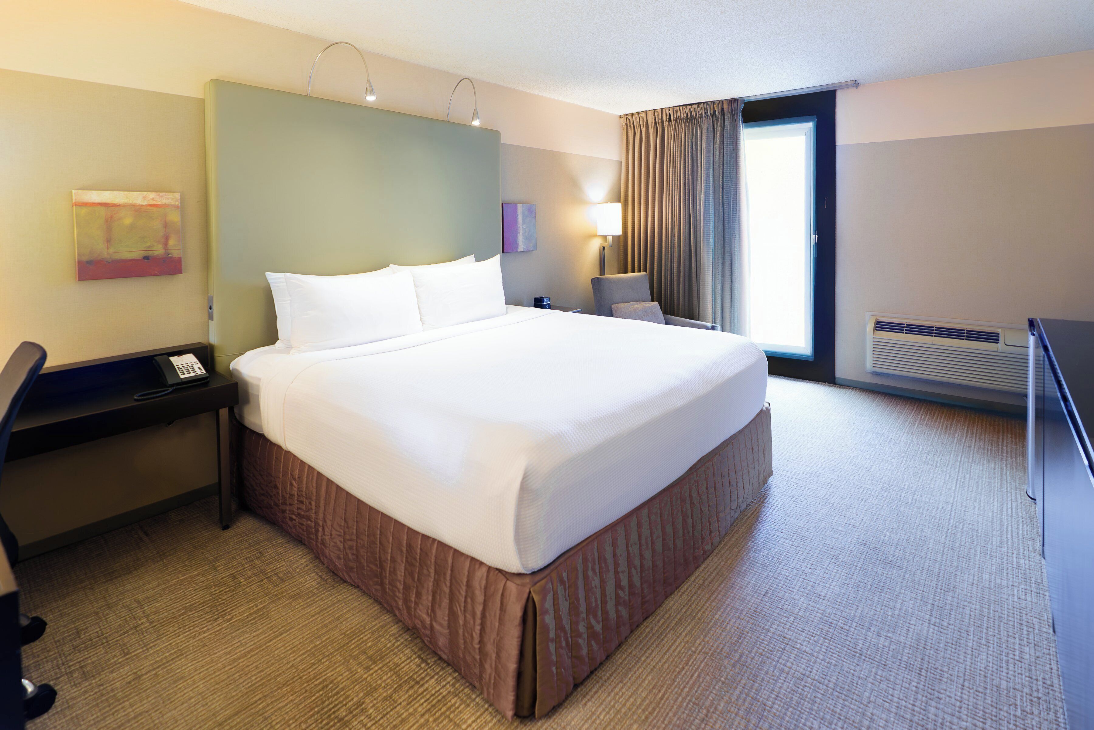 Une chambre d’hôtel avec un grand lit, un bureau, une chaise, une lampe de bureau et une fenêtre avec des rideaux.