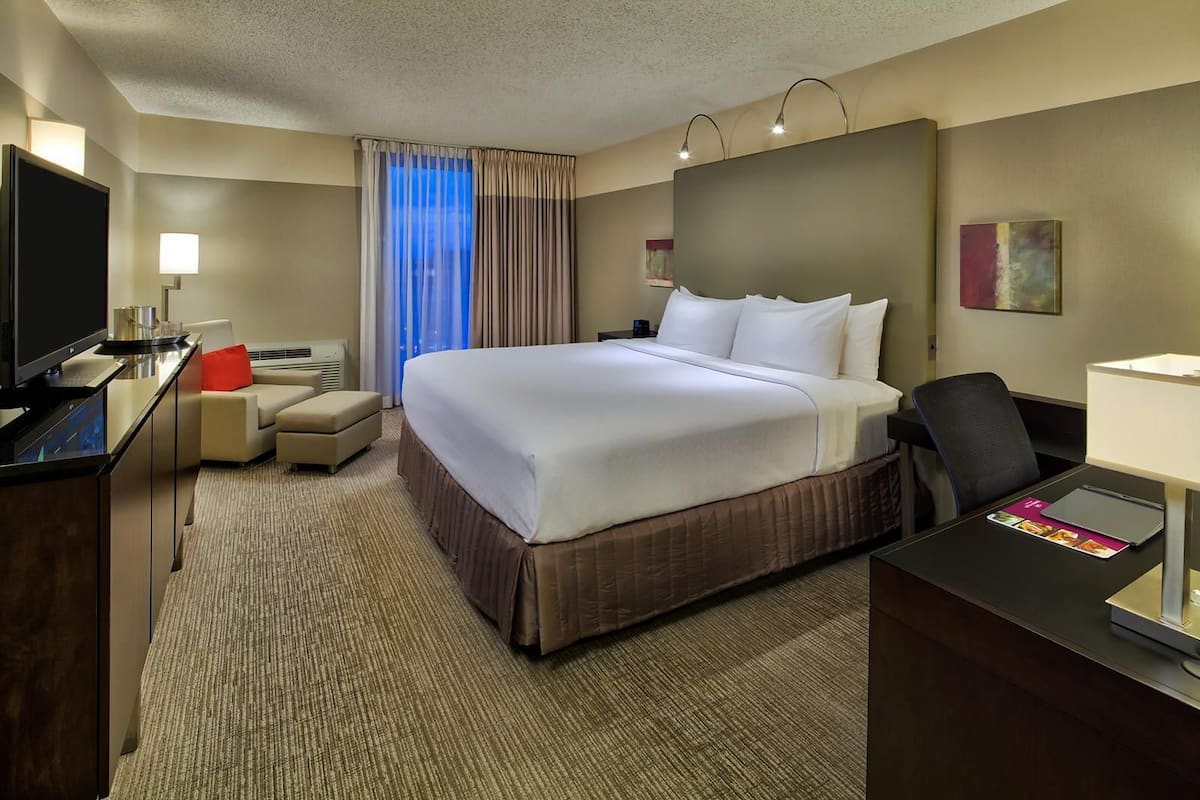A hotel room with a large bed, a desk with a computer, a TV, and a seating area with a chair and ottoman.