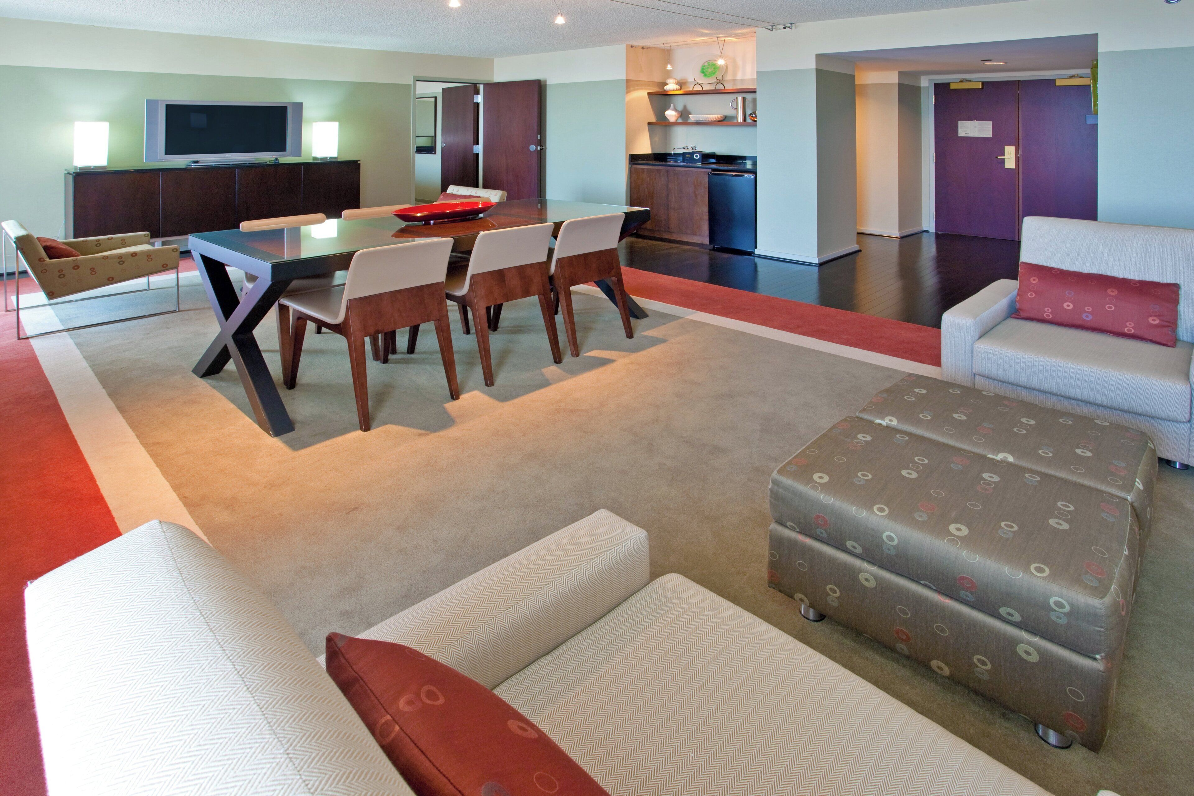 A modern living room with a dining table, chairs, a sofa, ottoman, and a kitchen area with cabinets and appliances.