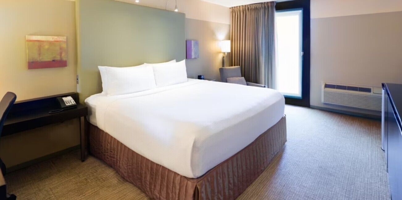 Ein Hotelzimmer mit einem großen Bett, einem Schreibtisch mit Telefon, einem Sessel, einer Lampe und einem fenster mit Vorhängen.