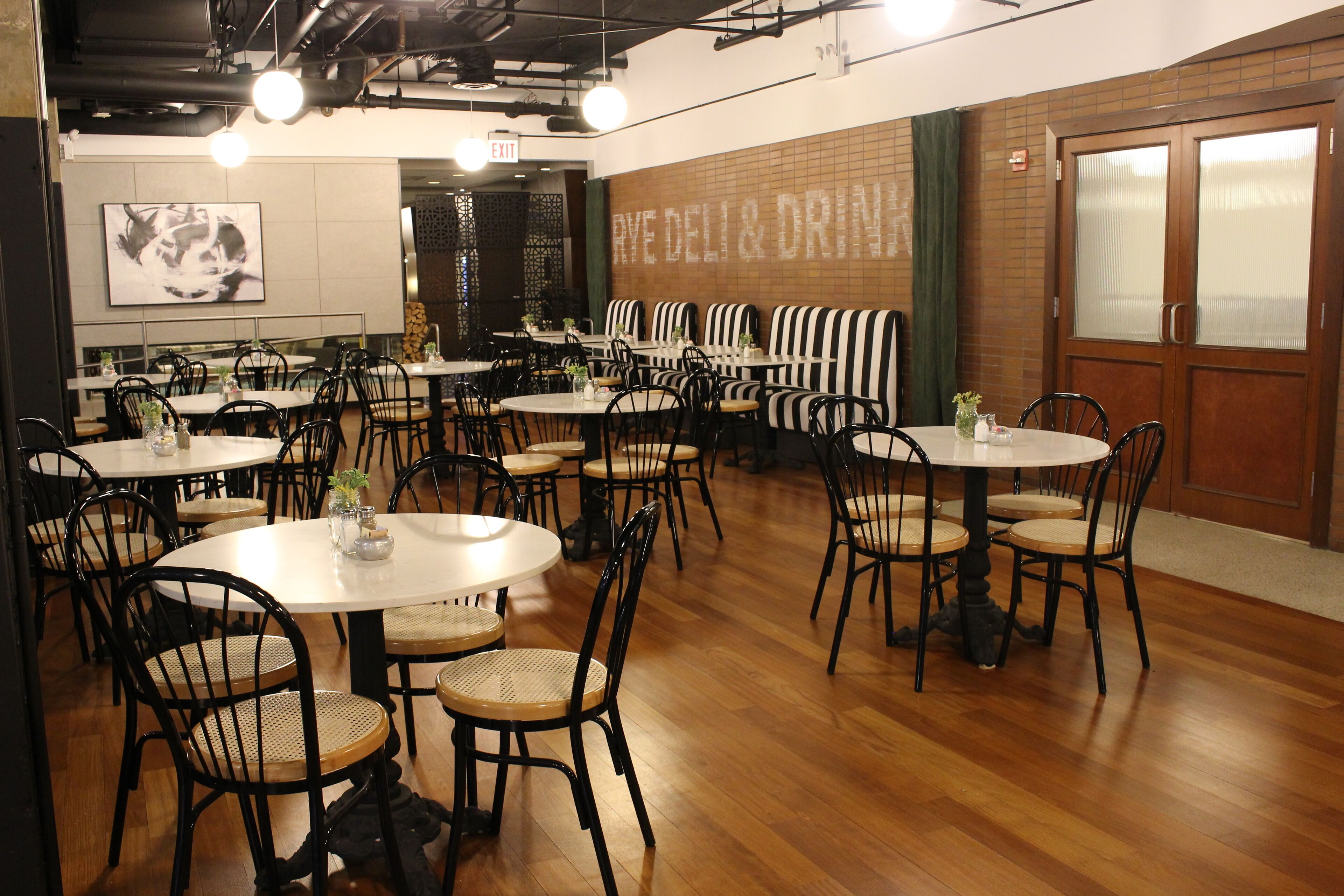 A restaurant with round tables and black chairs, a wall with a 'RVE DELI & DRINK' sign, and a wooden floor.