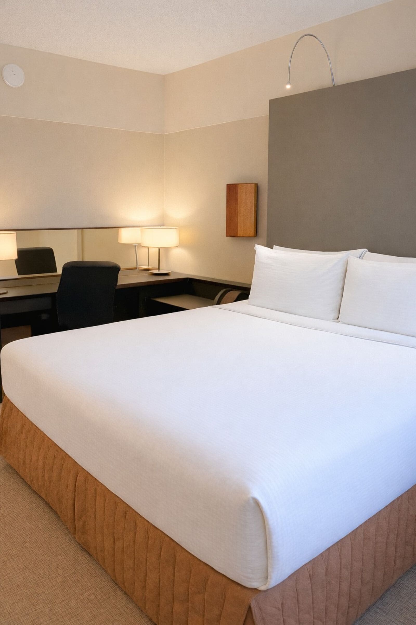 A hotel room with a large bed, a desk with a chair, a lamp, and a wall-mounted clock.
