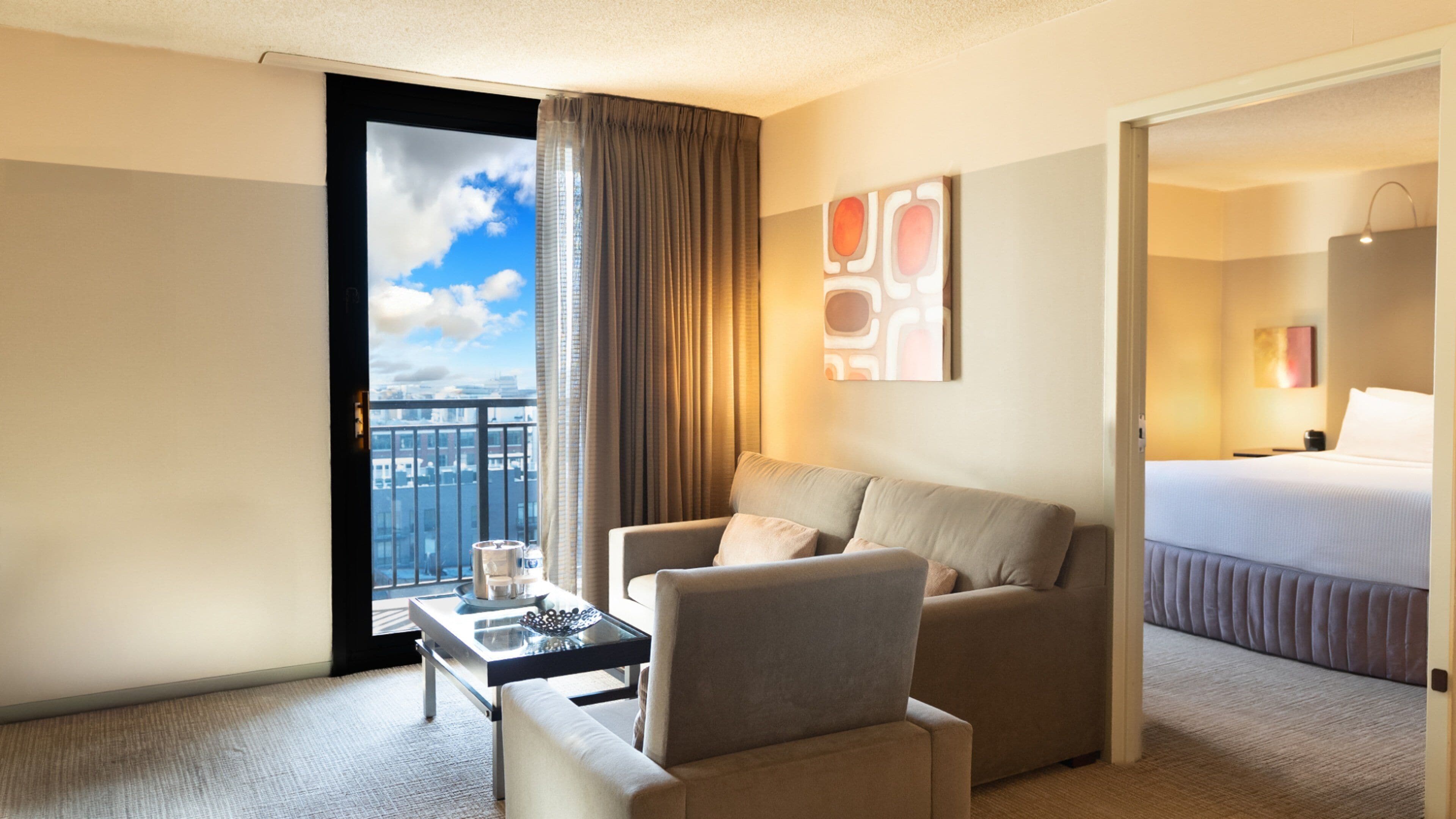 A hotel room with a sofa, a small table, a bed, and a view of the city through a window.