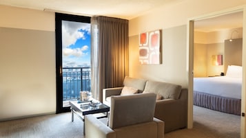 A hotel room with a sofa, a small table, a bed, and a view of the city through a window.