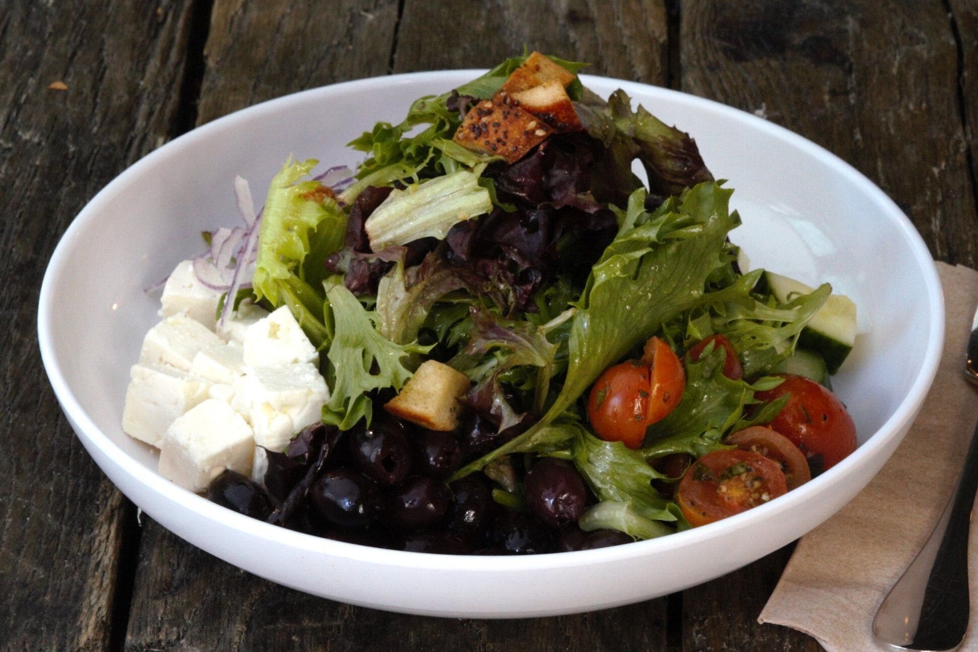 A white bowl containing a salad with mixed greens, feta cheese, cherry tomatoes, and cucumber slices.