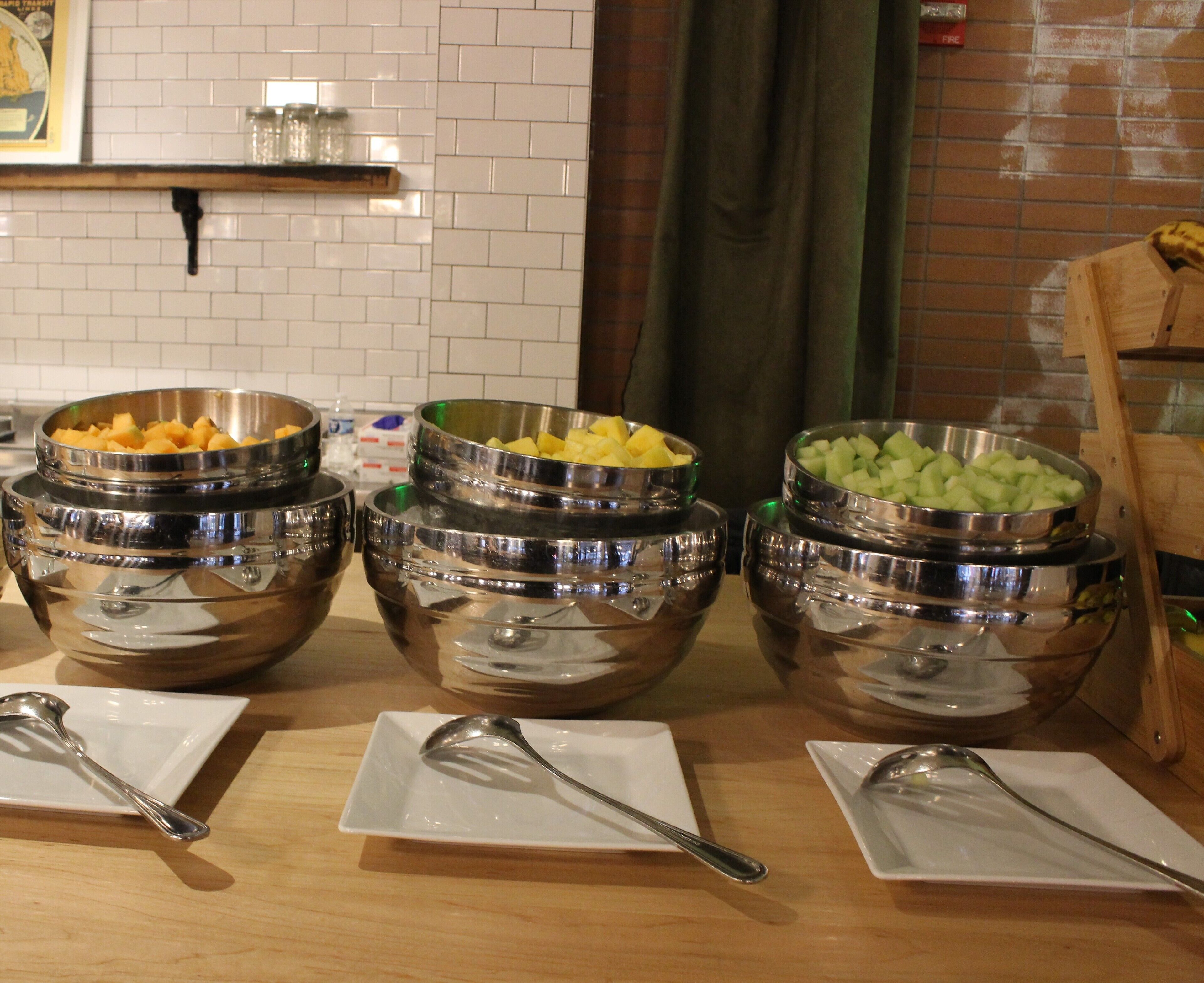 A buffet table with multiple stainless steel bowls containing various fruits and vegetables, accompanied by serving spoons on white plates.