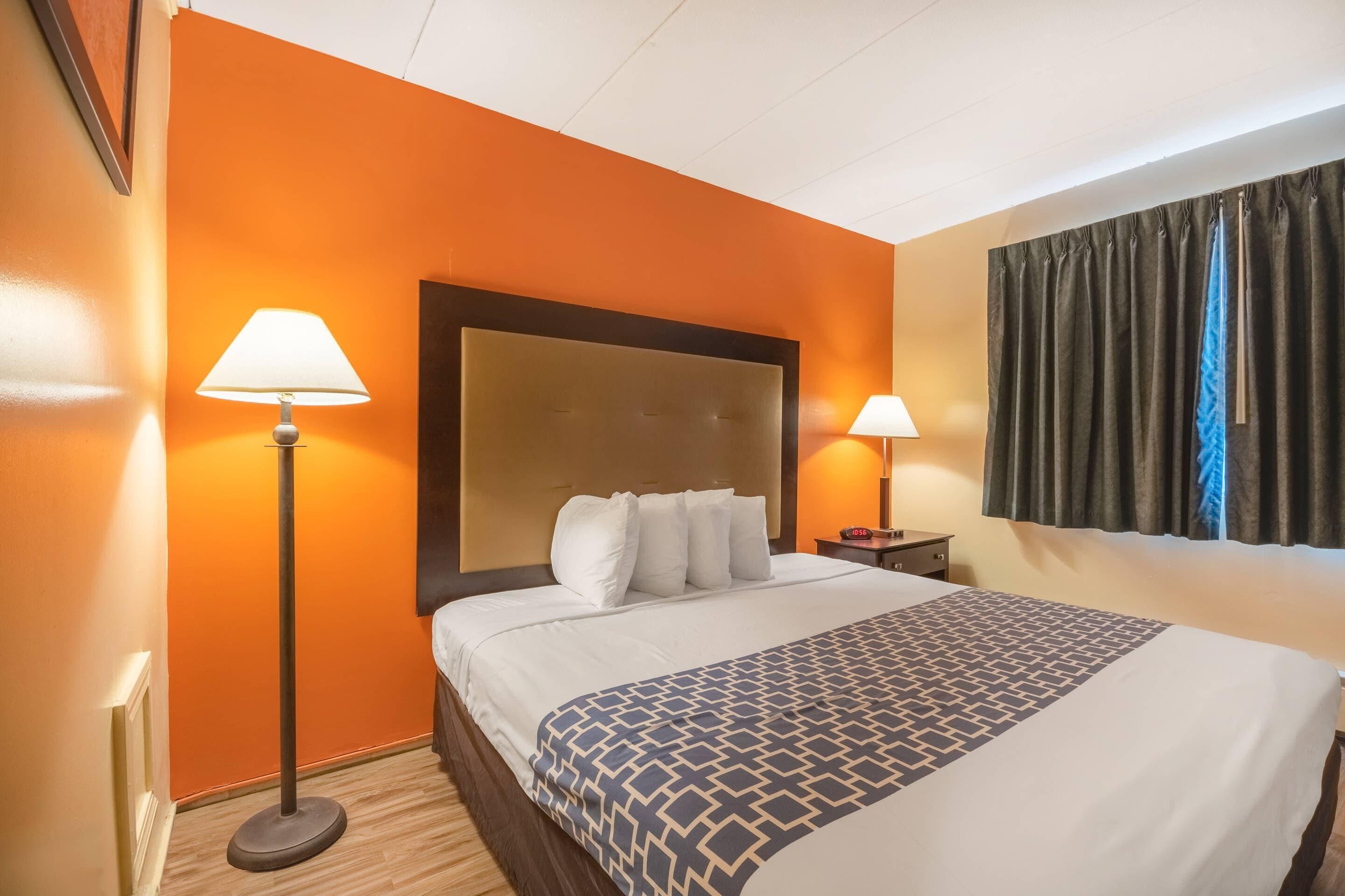 A hotel room with an orange wall, a large bed with a patterned bedspread, two bedside lamps, a nightstand, and a window with curtains.