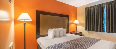A hotel room with an orange wall, a large bed with a patterned bedspread, two bedside lamps, a nightstand, and a window with curtains.