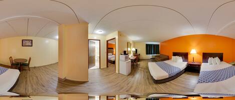 Vista panorámica de una habitación de hotel con techo curvo, un comedor con mesa y sillas, una kitchenette y dos camas con cabeceros iguales.