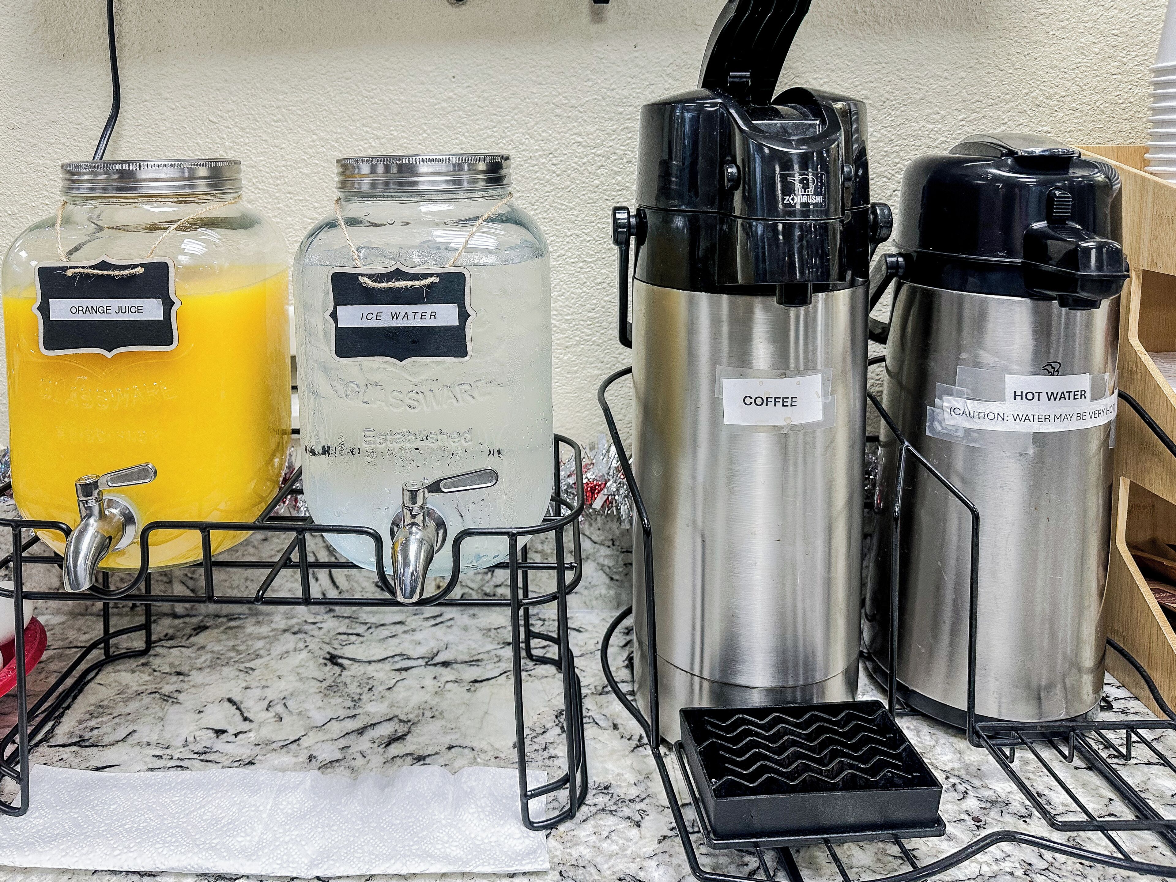 Een kitchenette met een koffiezetapparaat, een warmwaterkraan en twee glazen potten met 'sinaasappelsap' en 'ijs water'.
