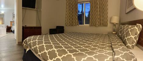 A bedroom with a bed, a desk, a chair, a window with curtains, and a TV mounted on the wall.