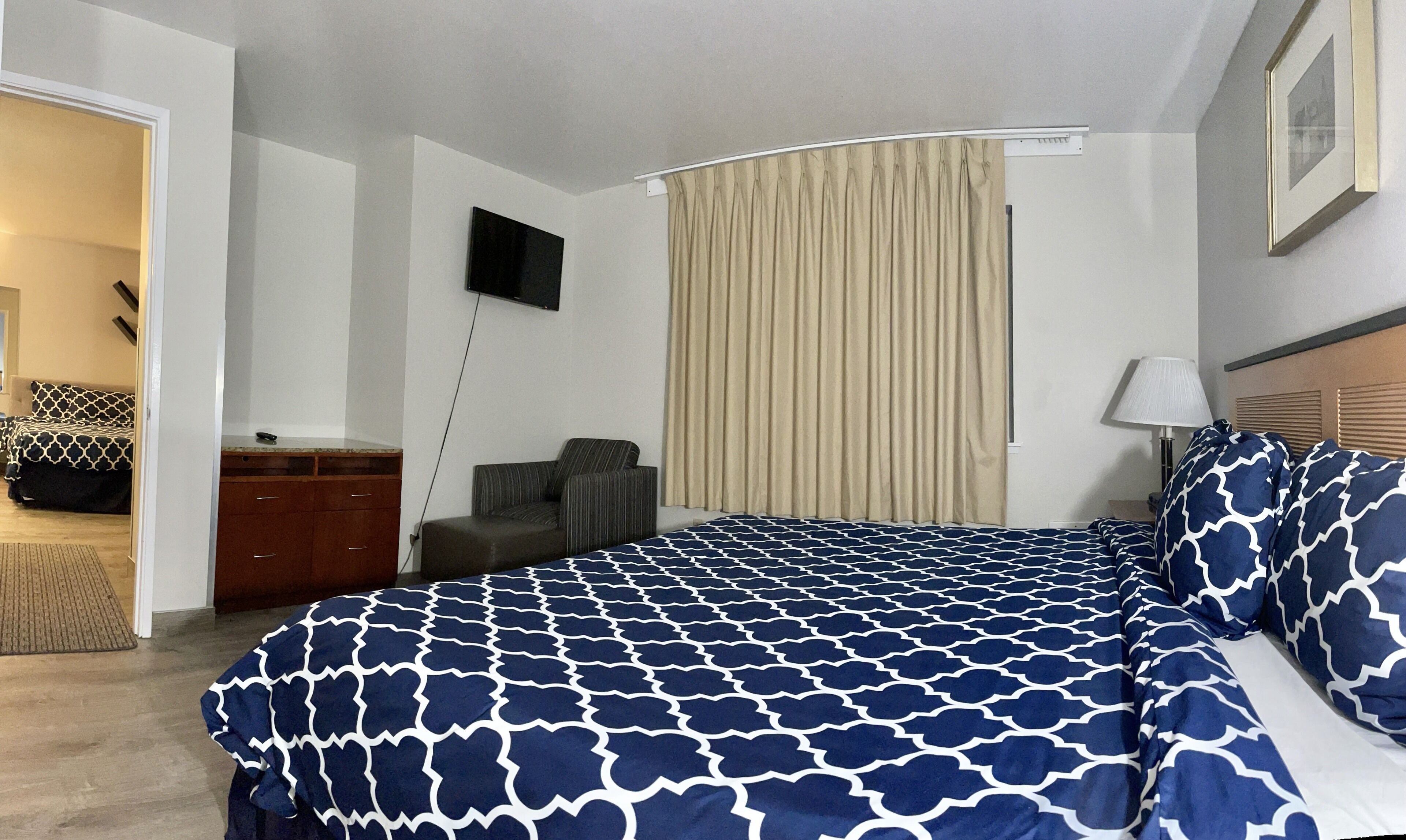 A hotel room with a large bed, a TV mounted on the wall, a desk with a chair, and a window with curtains.