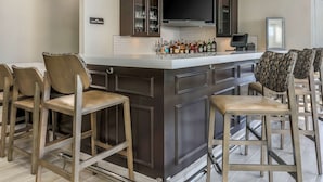 A modern kitchen with a bar area, featuring dark cabinetry, a large flat-screen TV, and a variety of bottles on the counter. - Hilton Garden Inn Tulsa South (Tulsa)
