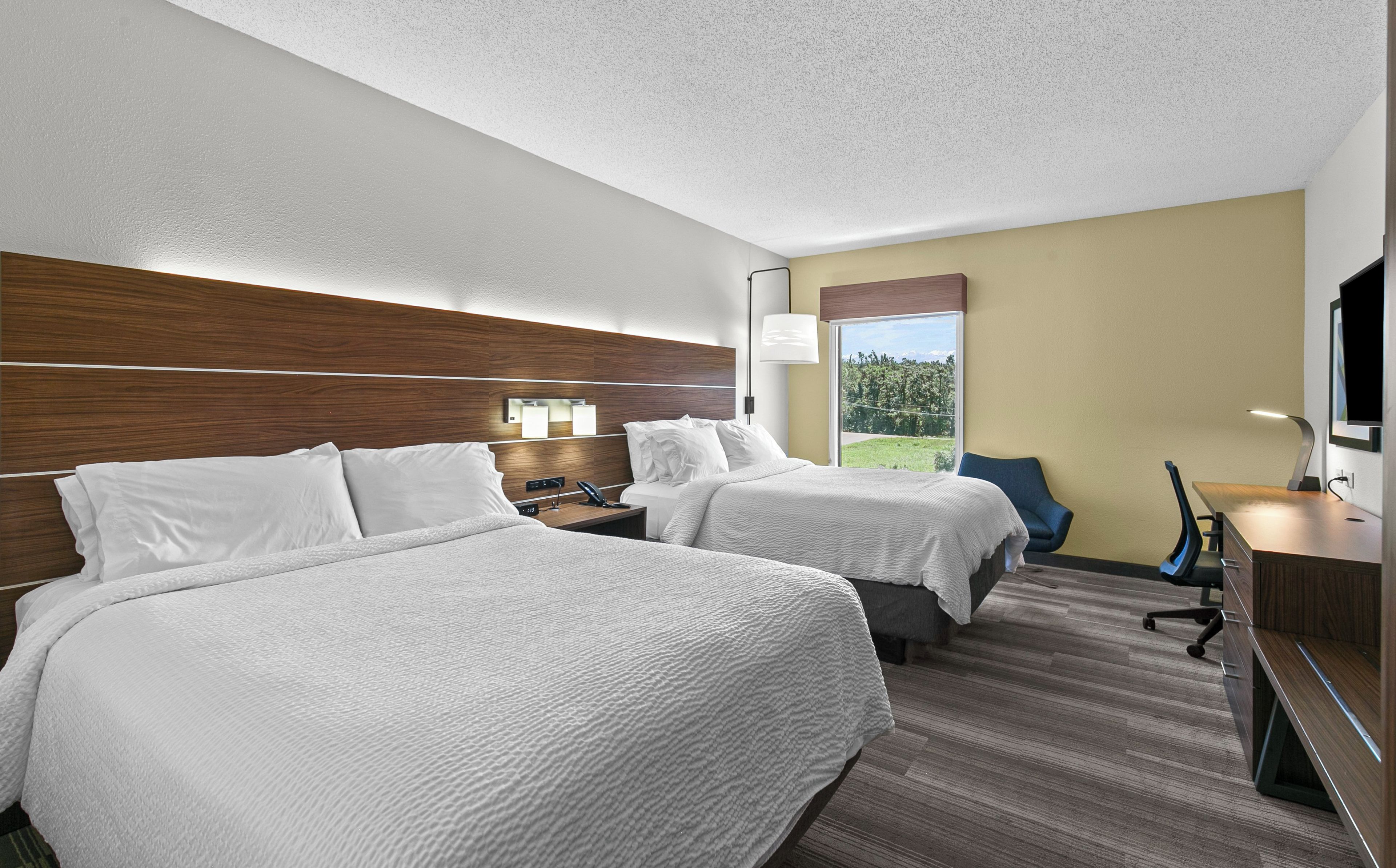 A hotel room with two beds, a desk, a chair, a TV, and a window with a view of greenery.
