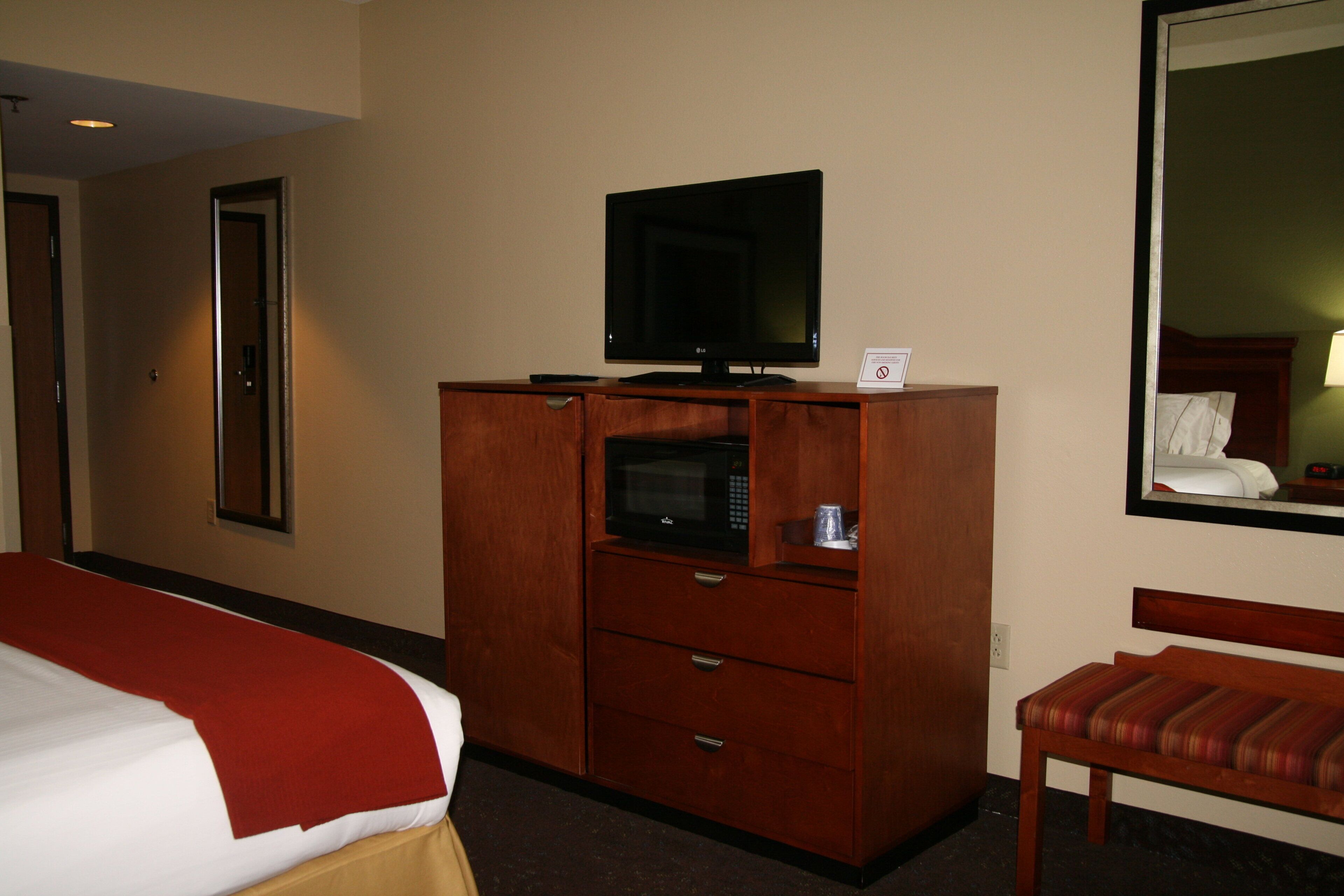 A hotel room with a bed, a wooden dresser with a television and microwave, a chair, and a mirror reflecting a bed and a nightstand.