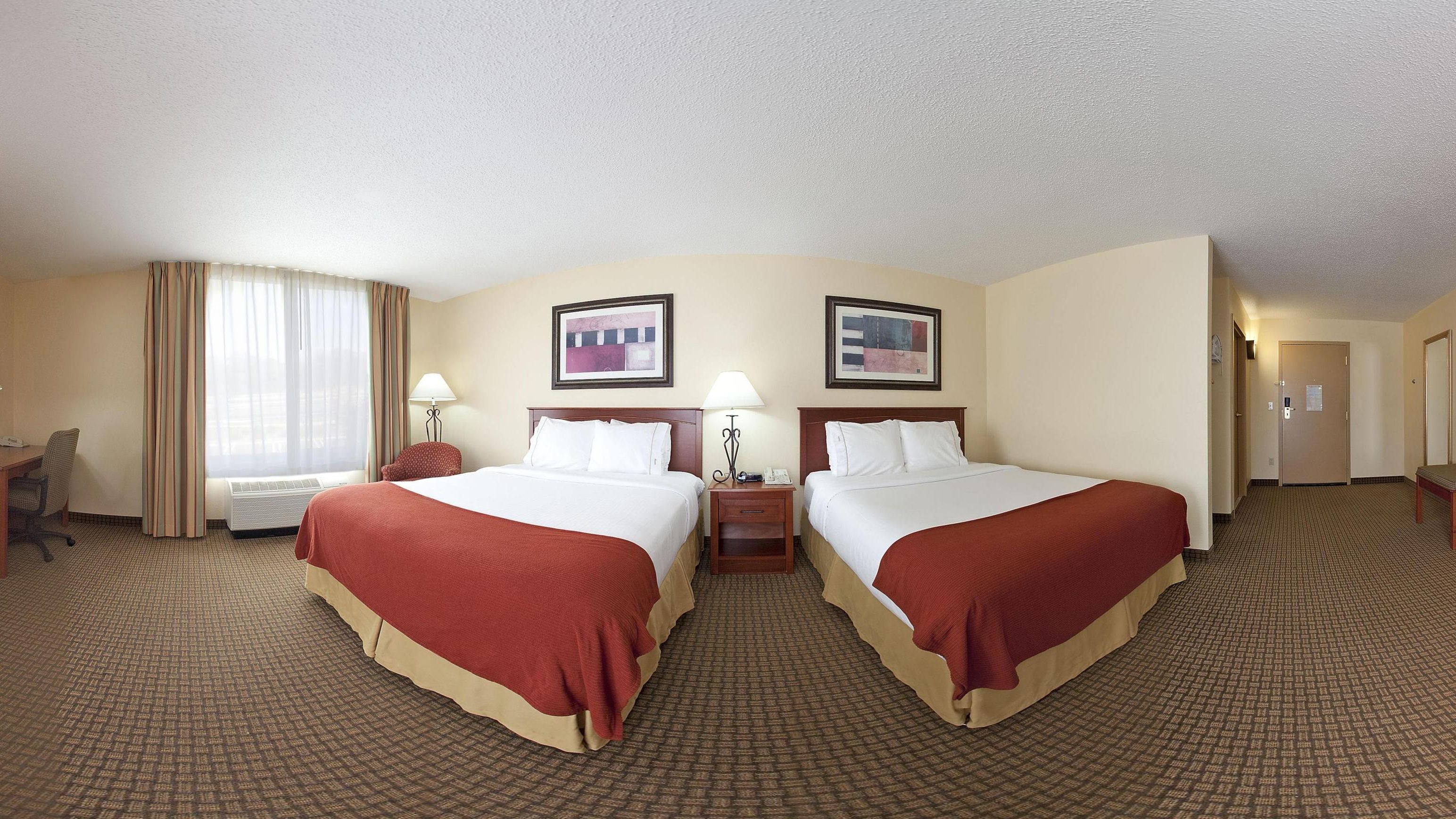 A hotel room with two beds, a desk, a chair, a lamp, and a window with curtains.