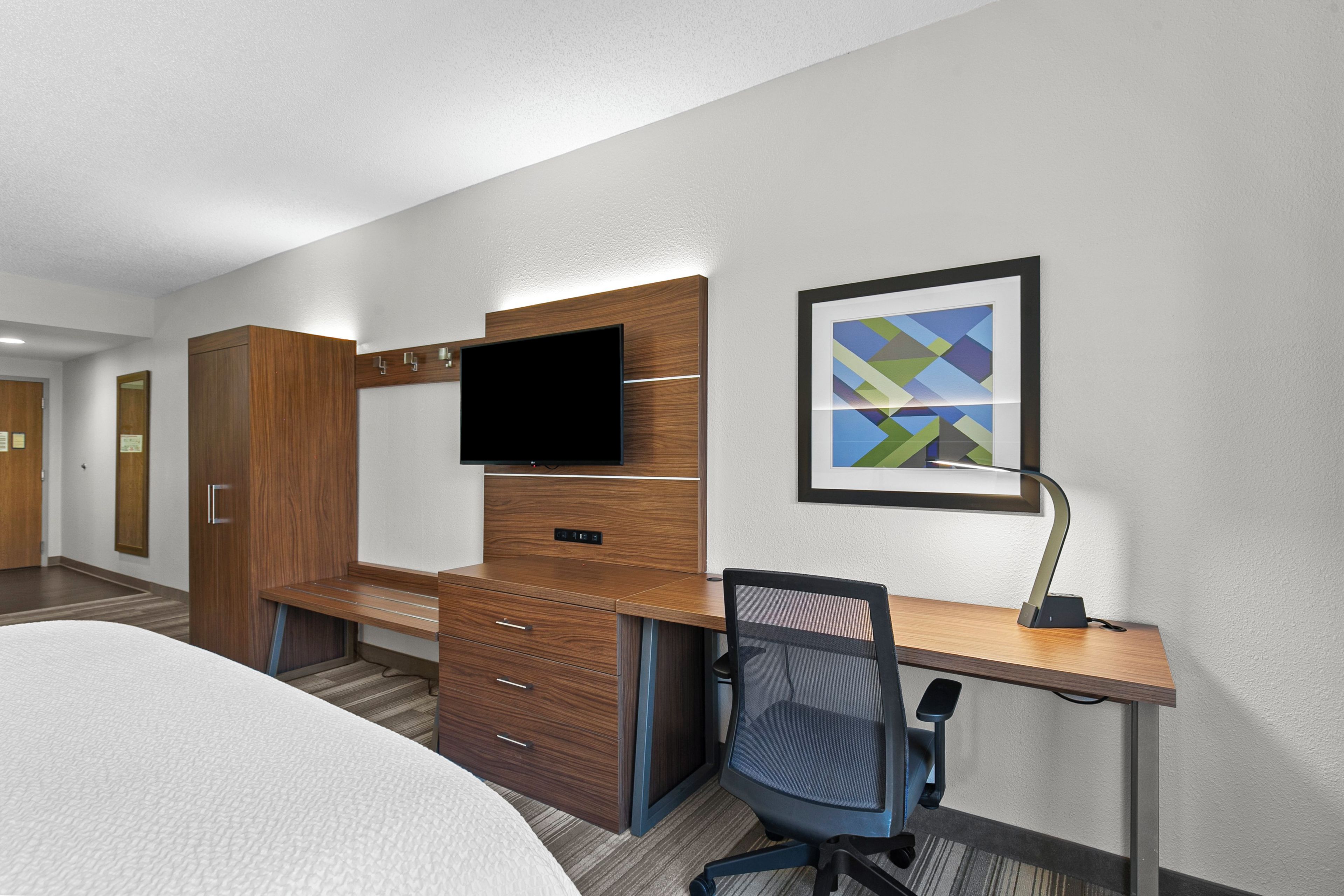 Ein Hotelzimmer mit einem Bett, einem Schreibtisch mit Stuhl, einem Fernseher, einem gerahmten abstrakten Gemälde und einem Kleiderschrank.