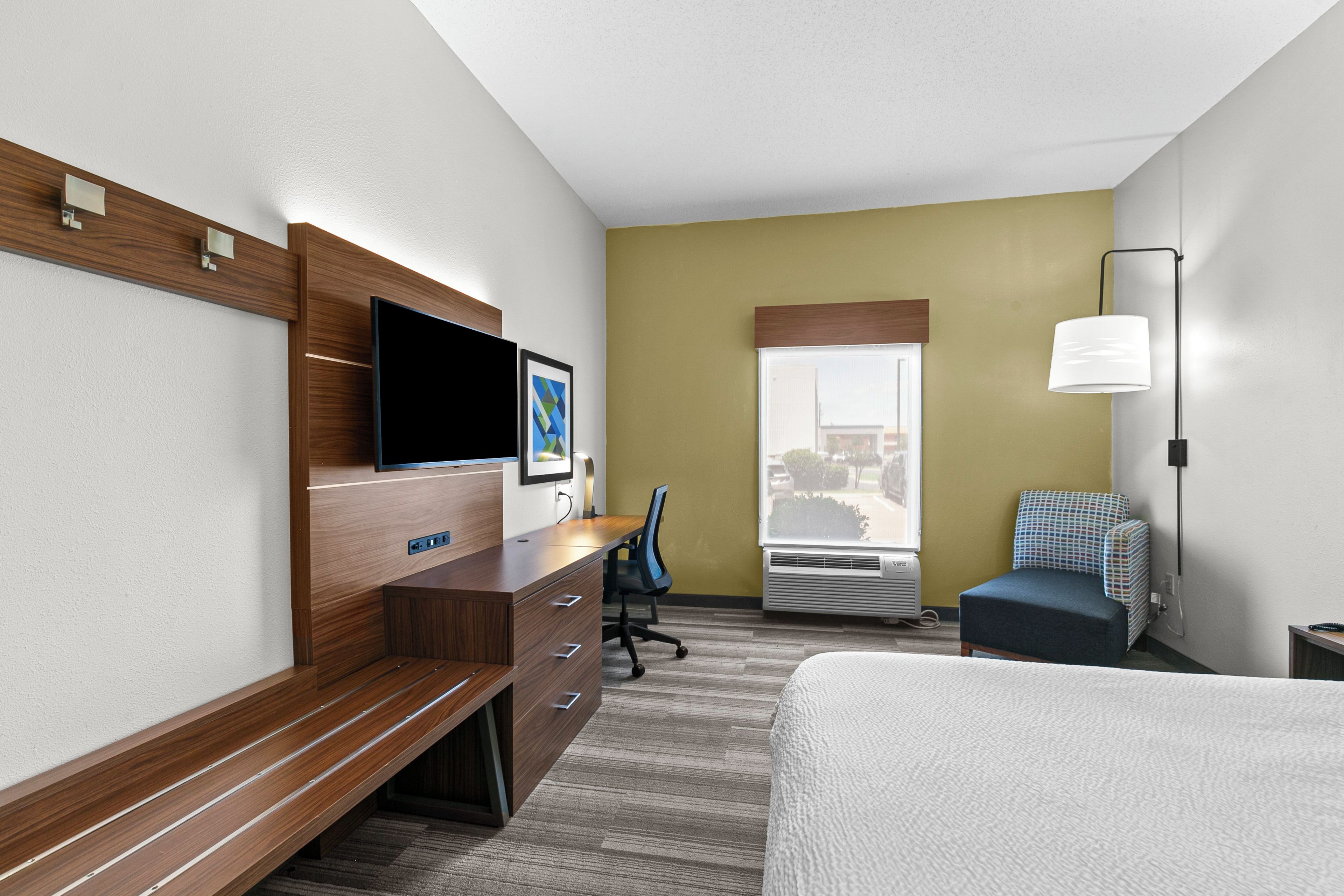Habitación de hotel con cama, escritorio con silla, televisión, ventana y lámpara.