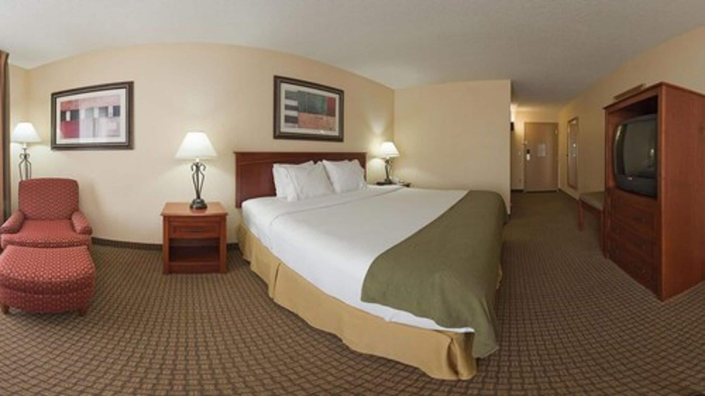 A hotel room with a large bed, two bedside lamps, a chair, a TV, and framed pictures on the walls.