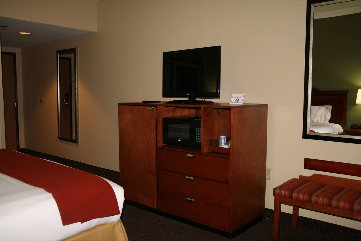 A hotel room with a bed, a wooden dresser with a television and microwave, a mirror, and a wooden bench.