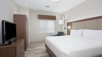 A hotel room with a bed, a television on a wooden cabinet, a window with blinds, a bedside table, and a lamp.