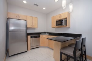 A modern kitchen with a stainless steel refrigerator, dishwasher, wooden cabinets, a black countertop, and a dining area with a chair.