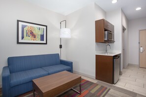 A modern room with a blue sofa, a wooden coffee table, a kitchen area with a microwave and sink, and a framed abstract painting on the wall. - Holiday Inn Express & Suites Amarillo by IHG (Amarillo)