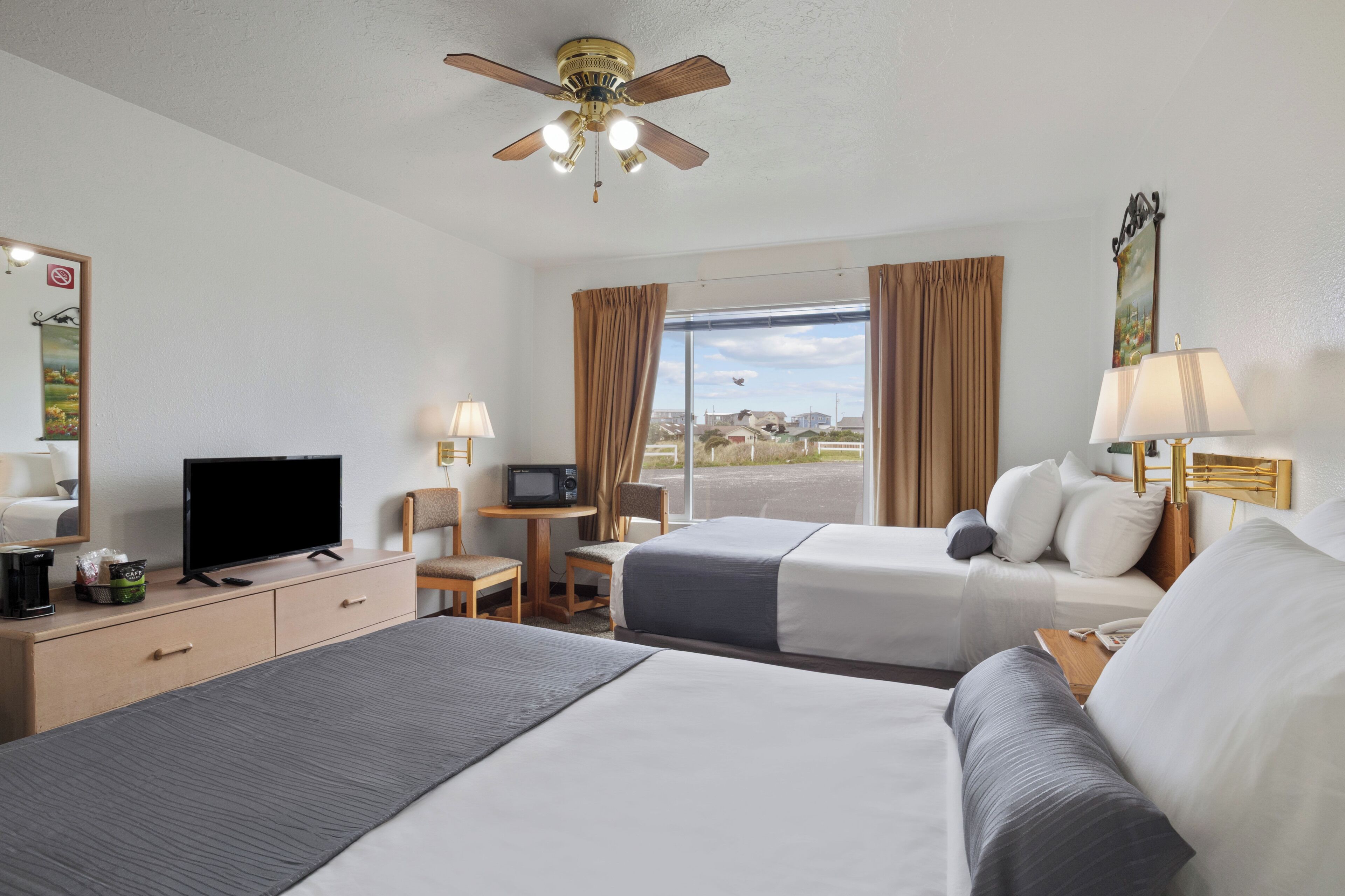 A hotel room with two beds, a television, a desk, a chair, a ceiling fan, and a window with curtains.