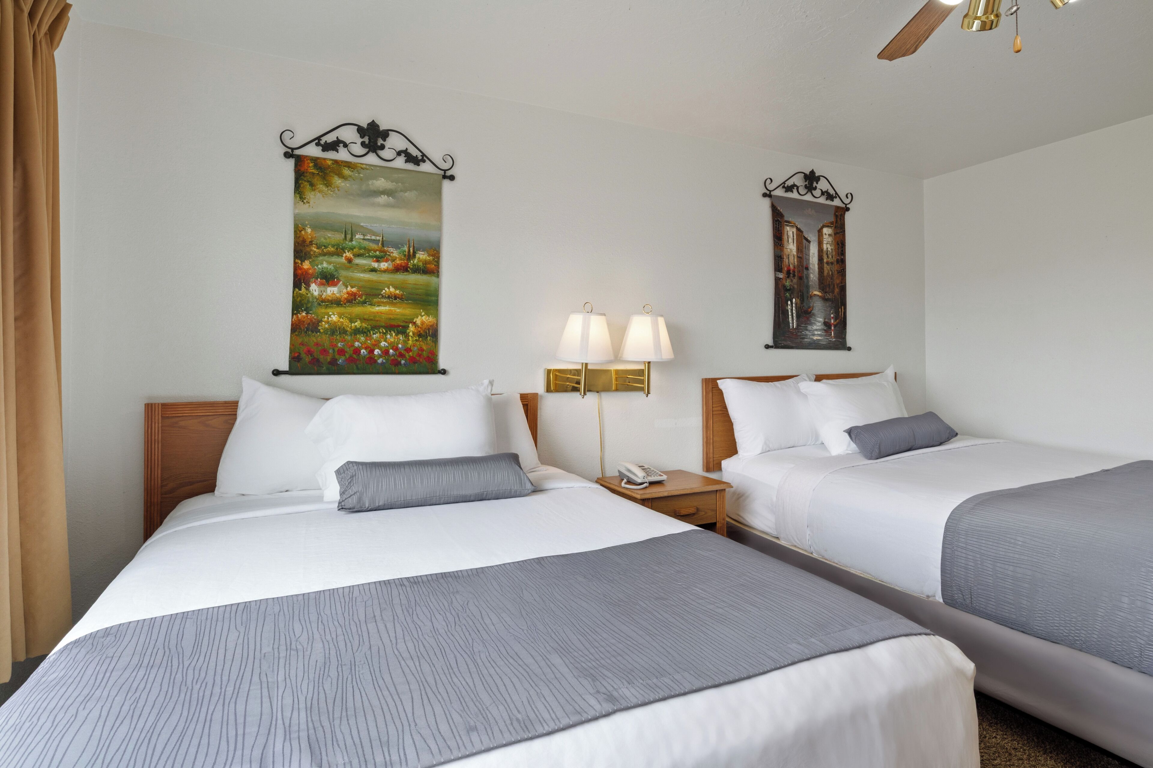 A hotel room with two beds, a nightstand with a lamp, and two paintings on the walls.