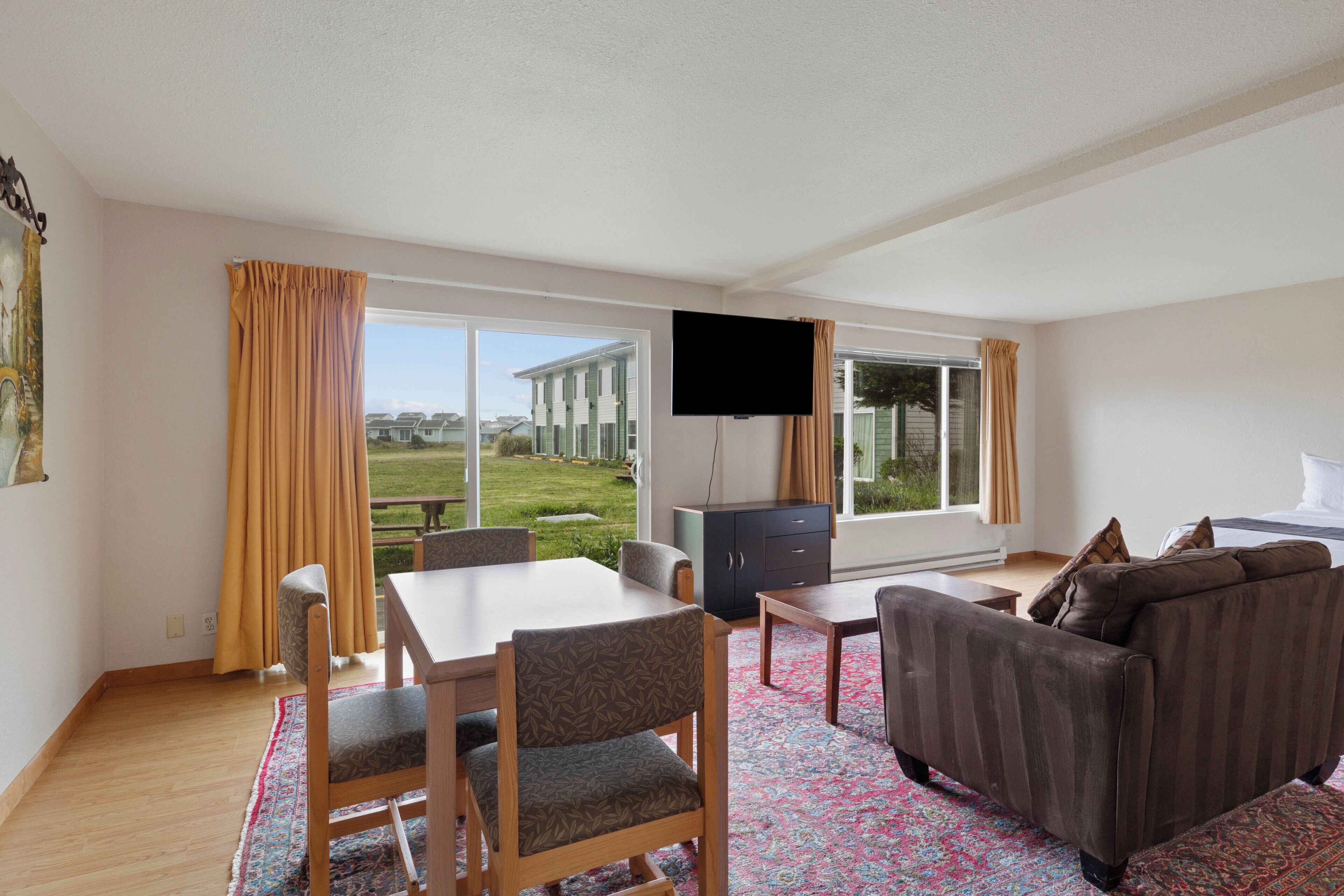 A modern living room with a dining table, sofa, and TV. Large windows offer a view of a building and greenery.