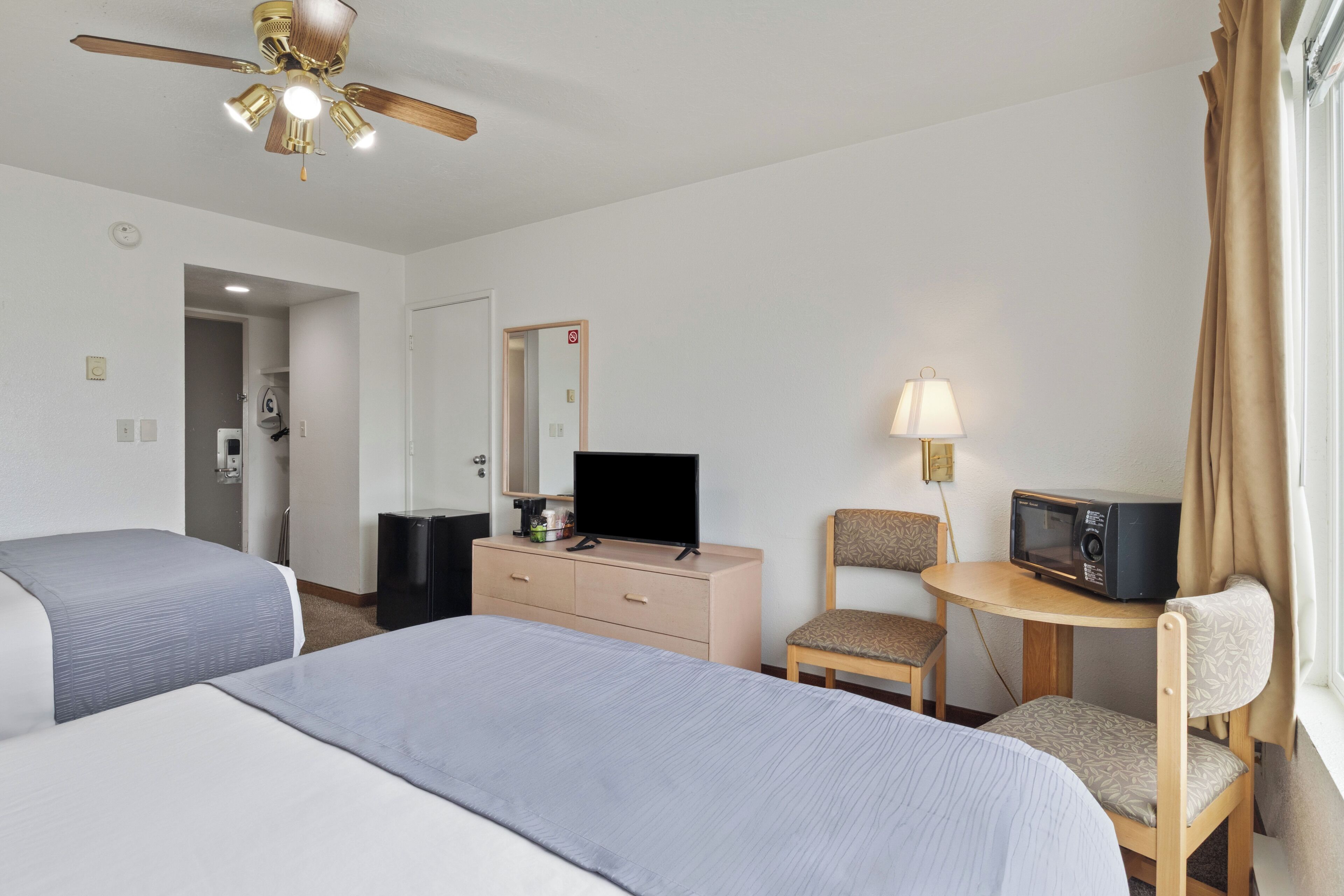 Habitación de hotel con dos camas, ventilador de techo, televisión, microondas, una silla y una lámpara.