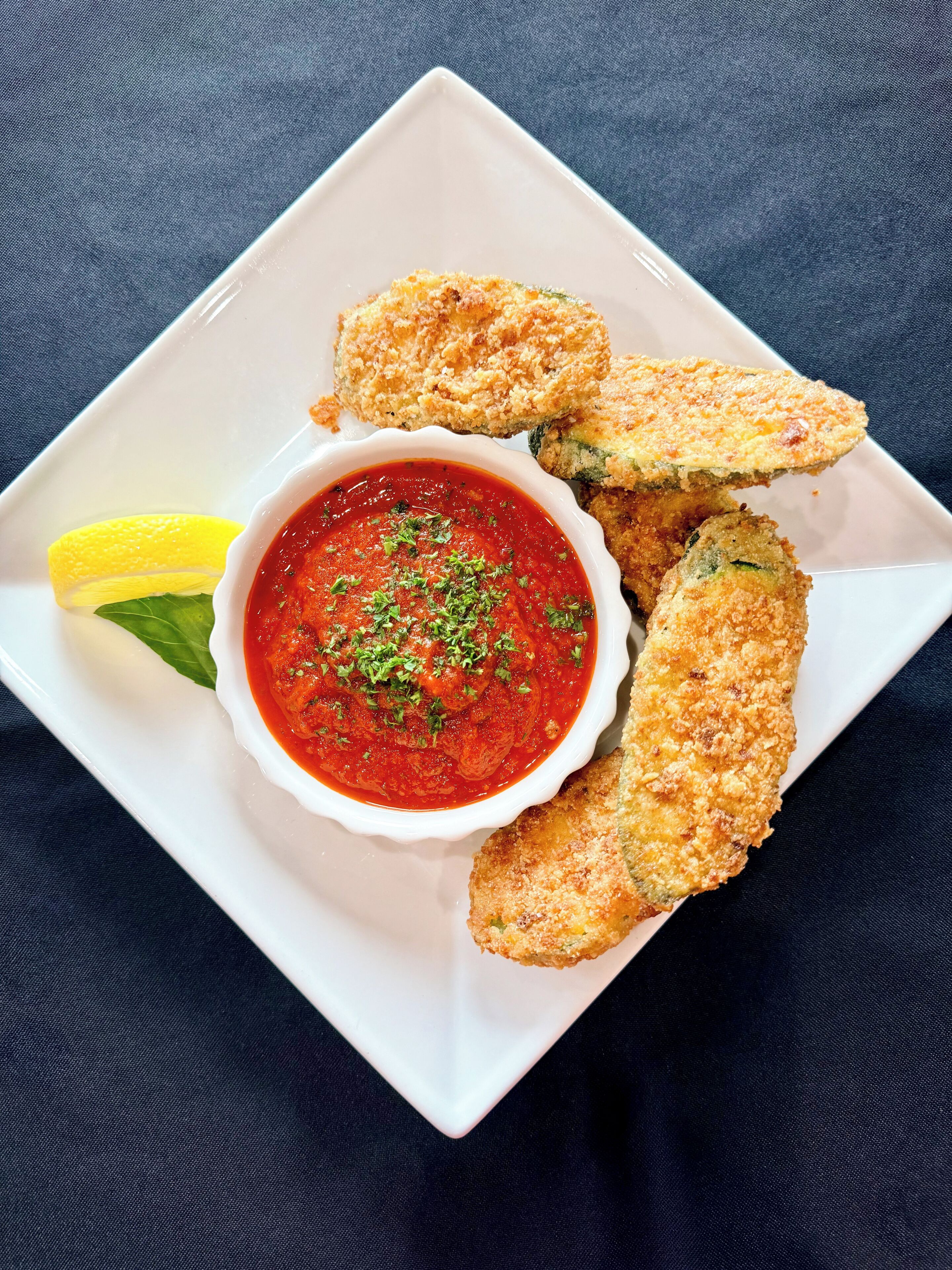 A white plate with breaded zucchini slices, a lemon wedge, and a bowl of red sauce garnished with herbs.